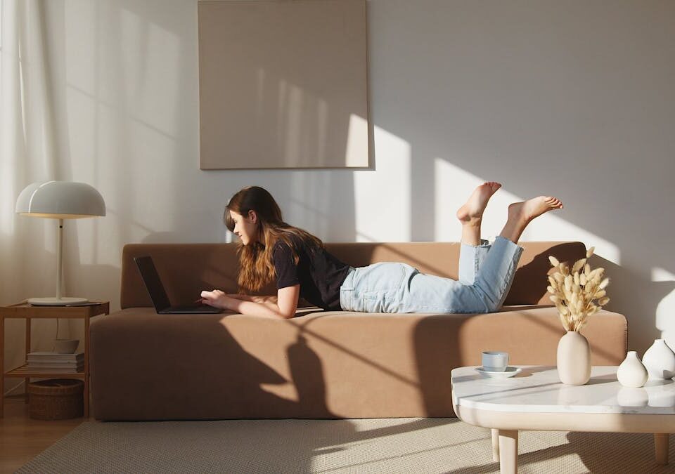 Side view full body young lady in casual outfit lying on comfortable couch and browsing netbook in cozy modern living room