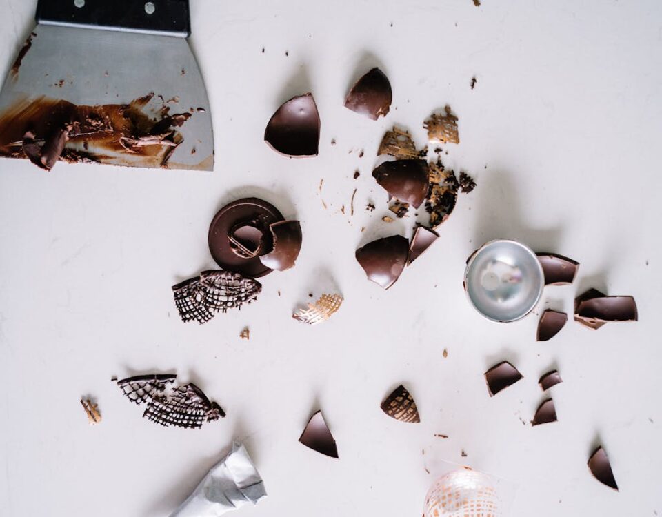 Flatlay of chocolate pieces and a scraper on a white surface, perfect for confectionery themes.