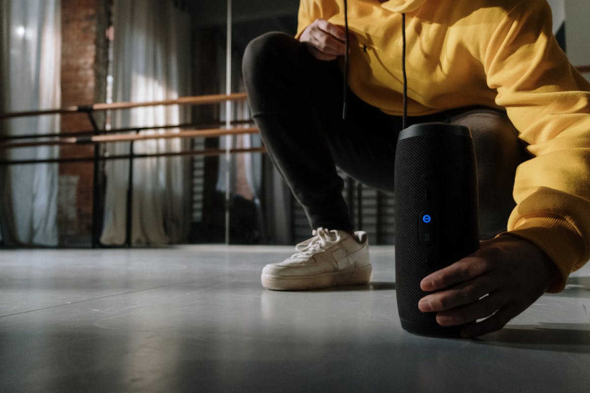 Close-up of a person in a yellow hoodie adjusting a wireless speaker on the dance studio floor.