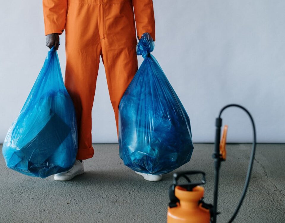 Sanitation worker in orange overalls holding blue trash bags indoors for waste management.