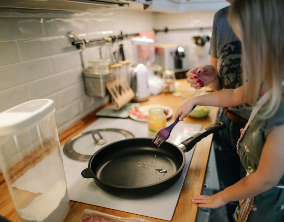 Young girl cooking with a frying pan under adult supervision in a modern kitchen.