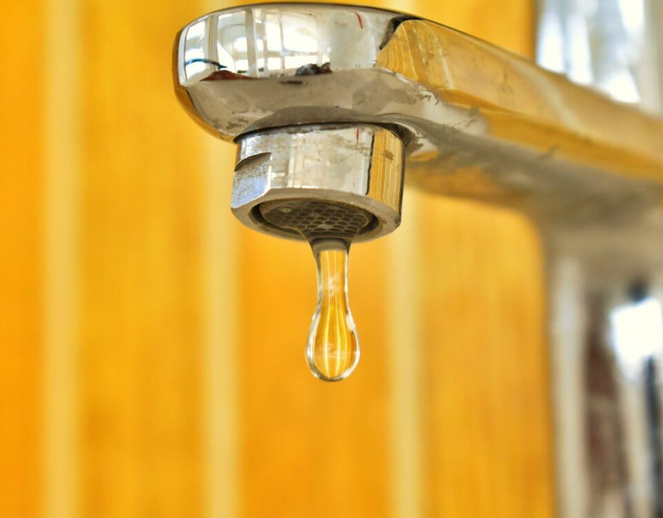 A close-up shot of a water droplet hanging from a stainless steel faucet, highlighting surface tension.