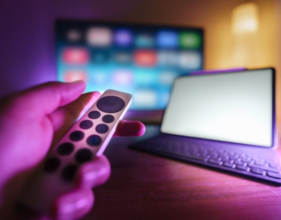 A hand using a remote to control a TV and laptop in a cozy room setting, perfect for relaxation.