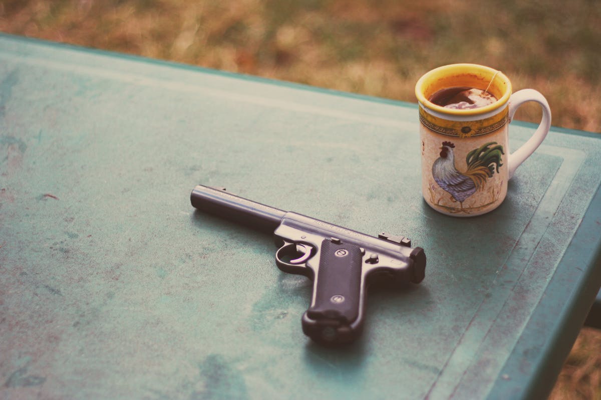 A handgun and rooster-themed mug with tea on a green outdoor table.