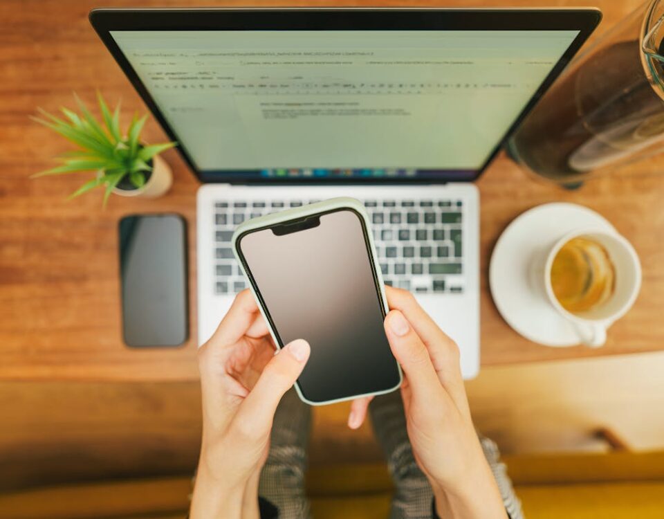 Hands holding a smartphone over a laptop with coffee in a cozy home office setup.