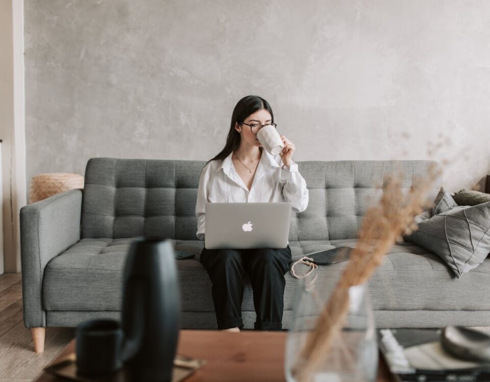 Woman sipping coffee while working on a laptop in a cozy home setting, highlighting remote work lifestyle.