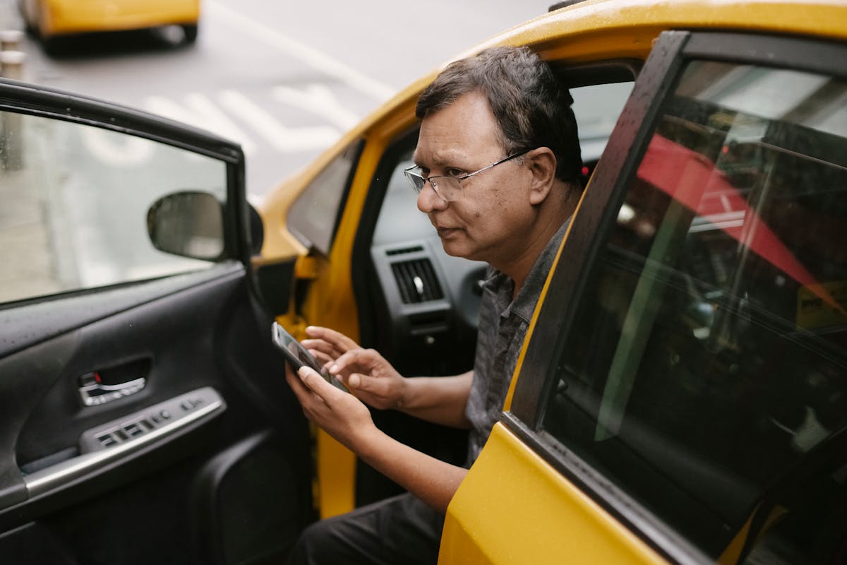 Crop Asian man sitting in parked yellow taxi car with open door and using smartphone while looking away