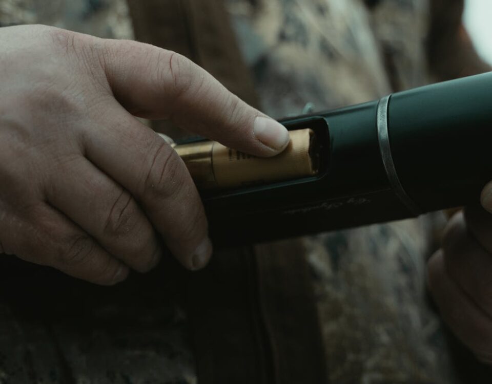 Close-up of a person inserting a bullet into a shotgun with camouflage attire in the background.