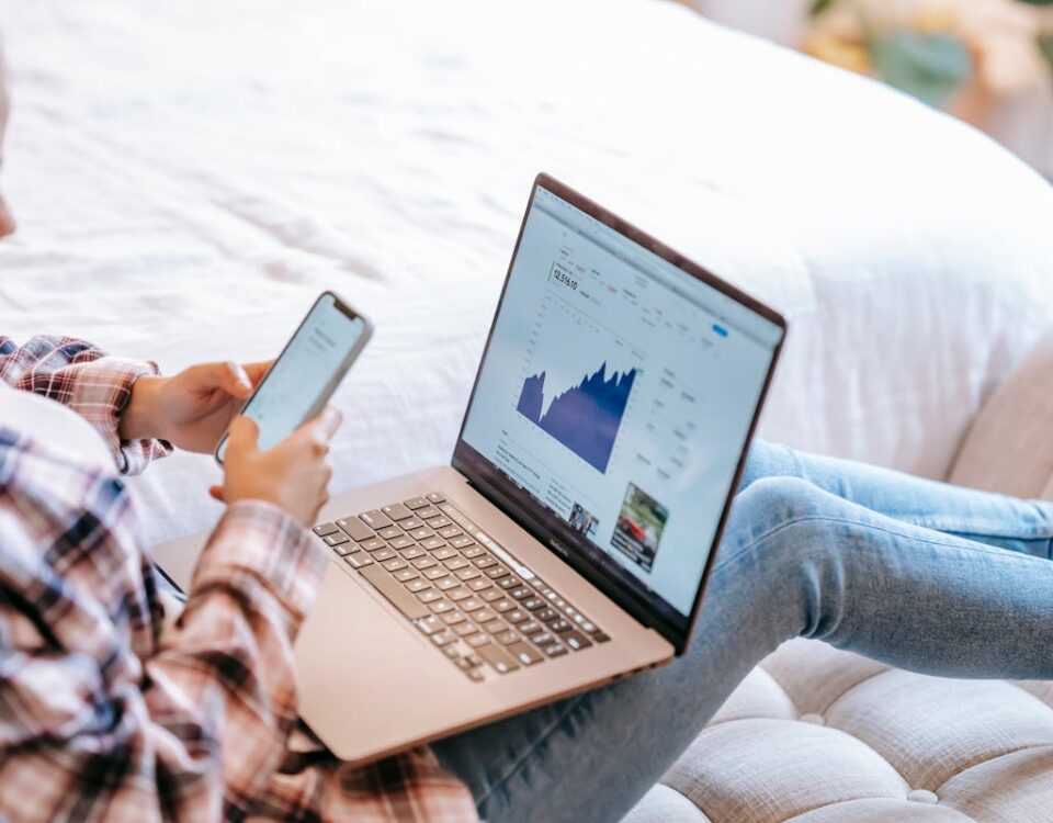 A woman using a smartphone and laptop to analyze stock market data from home.