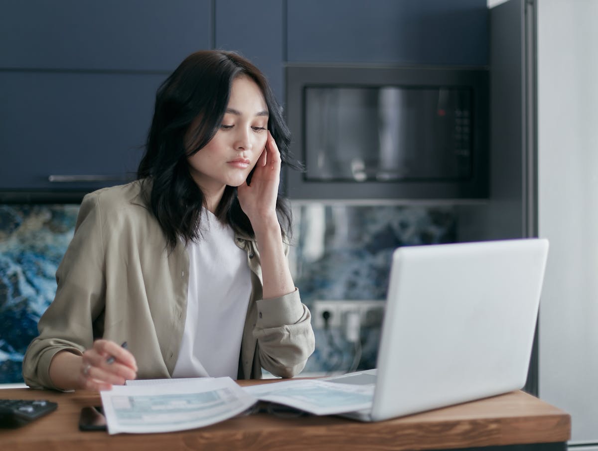 A young woman focused on her laptop, reading papers in a modern kitchen setting.