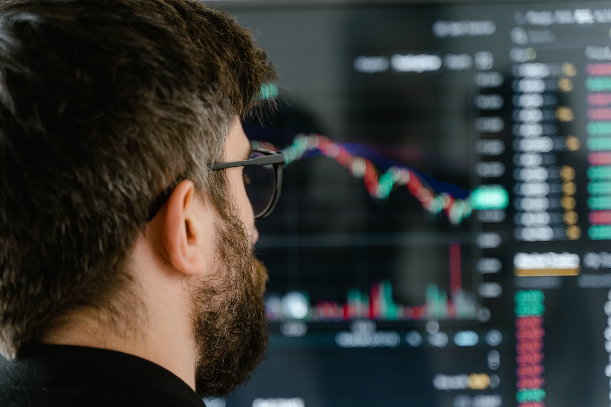 A bearded man with glasses examines trading charts on a computer screen, indicating financial analysis.