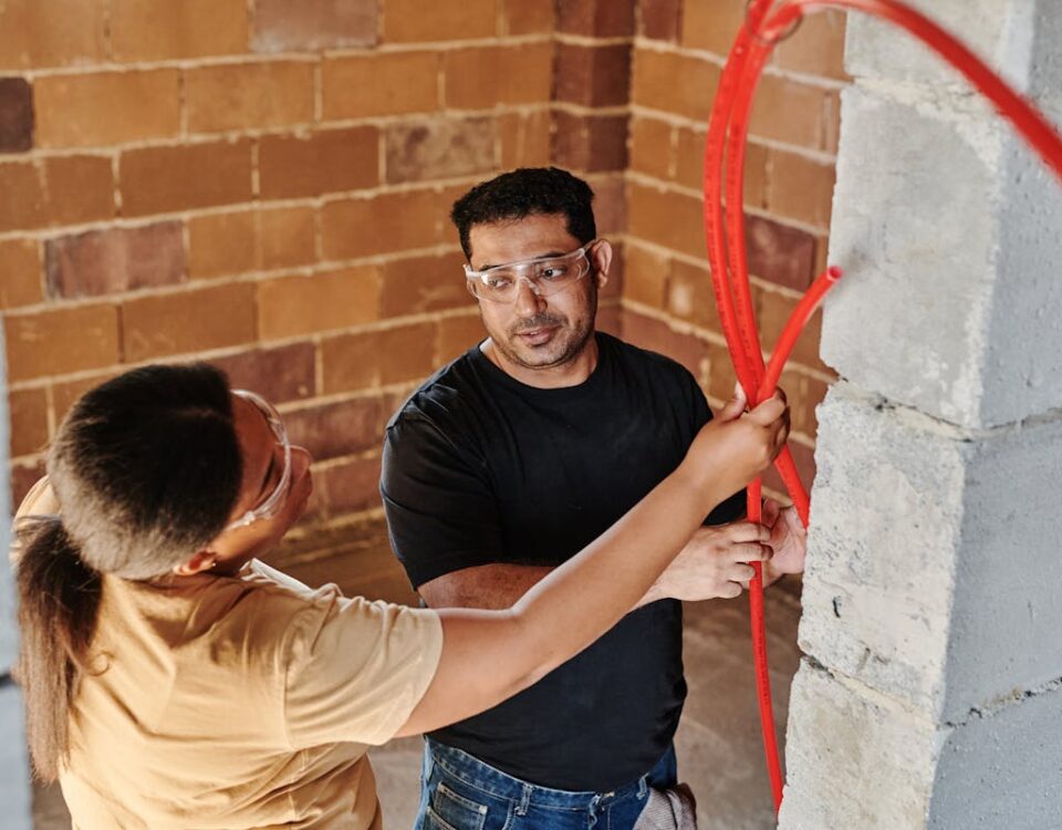 Two construction workers fitting red tubing on a brick wall at a construction site.