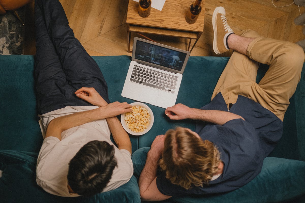 Two friends enjoying a movie on a laptop with popcorn from a cozy couch perspective.