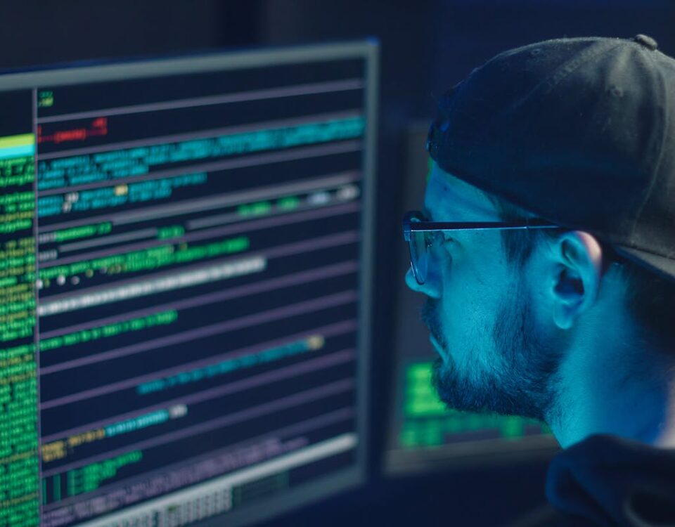 A cybersecurity expert inspecting lines of code on multiple monitors in a dimly lit office.